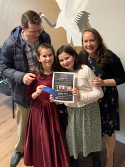 Family celebrating music education with a Bach Trio event poster, emphasizing community engagement in piano learning.